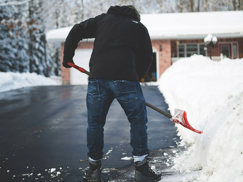 A man wearing a winter coat and hat shoveling heavy snow off a residential driveway